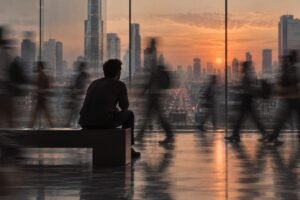 homem sentado imóvel observando a cidade ao entardecer, pessoas em movimento borrado ao redor, representando a pergunta se a solidão é a doença do século XXI