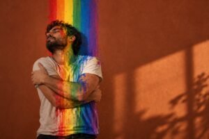Solidão gay depois dos 40: homem de barba e cabelo cacheado, de olhos fechados e braços cruzados em um abraço em si mesmo, com tinta nas cores do arco-íris escorrendo sobre o rosto e a camiseta, diante de uma parede terracota com sombra de janela