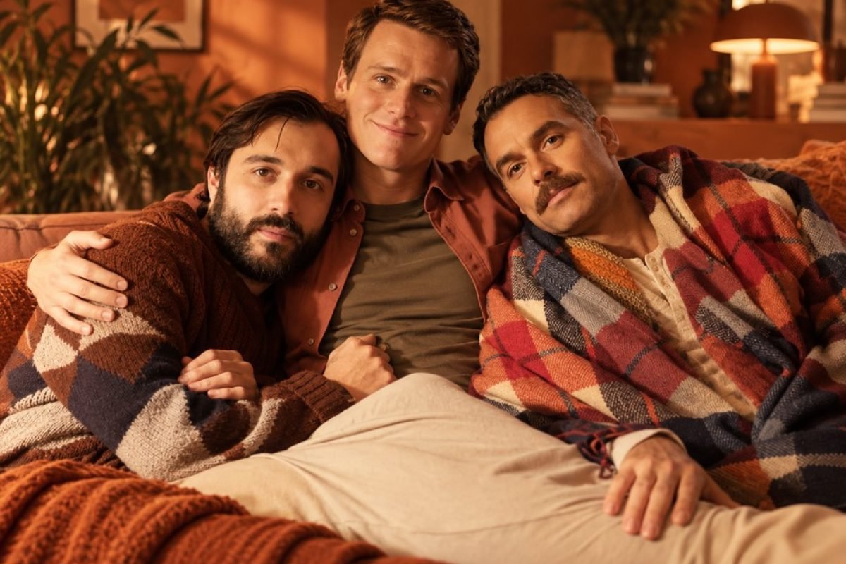 Solidão gay depois dos 40: três homens abraçados em um sofá, em uma sala acolhedora de tons terracota, o do meio de camisa avermelhada, o da esquerda de barba e suéter marrom, o da direita de bigode e coberto por uma manta xadrez