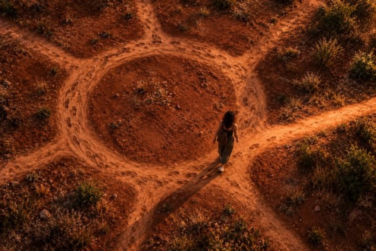 Trauma e repetição: vista aérea de uma pessoa caminhando por uma trilha circular marcada por muitas pegadas anteriores, com um dos pés levemente fora do caminho